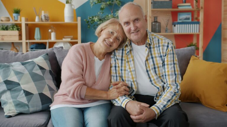 older couple seated on couch with the woman holding the man's arm and leaning into him