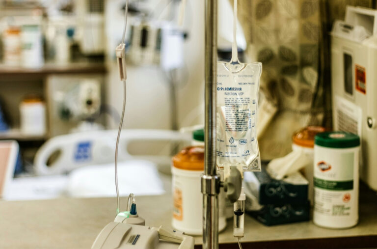 inside patient room at hospital, forefront shows IV bag on a pole, in the background is a table with medical supplies and a medial bed beyond the table