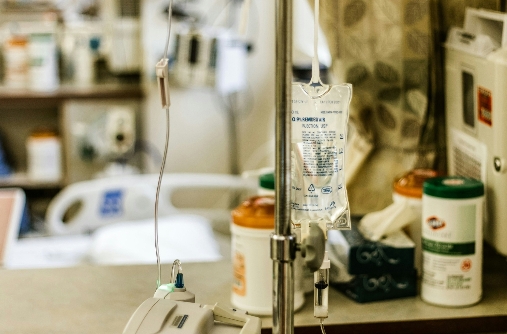 inside patient room at hospital, forefront shows IV bag on a pole, in the background is a table with medical supplies and a medial bed beyond the table